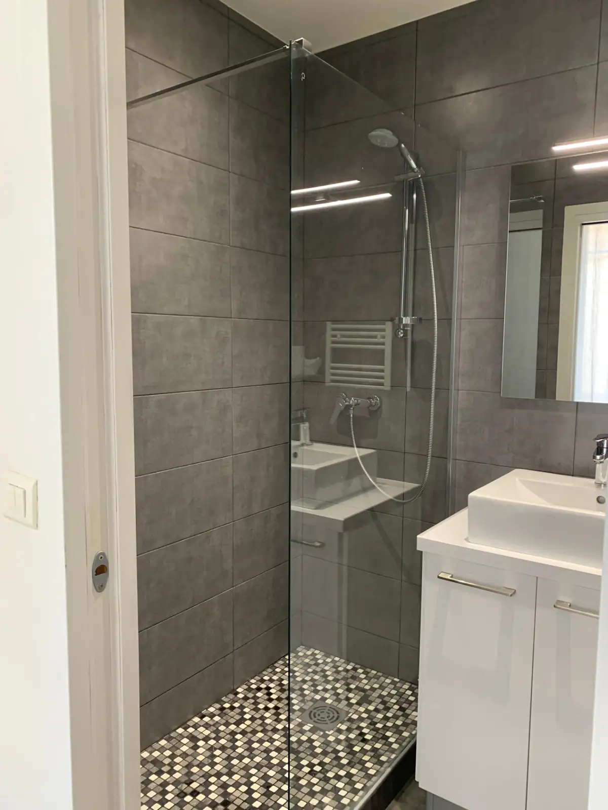 Glass walk-in shower over a mosaic floor beside a designer basin